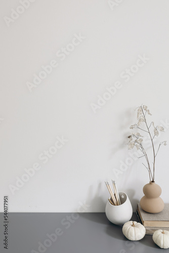 Artistic wokspace, still life. Pencils in ceramic holder. Vase with dry lunaria flowers, litttle white pumpkins, books. Grey table. Creative table background. Autumn, winter home office decor, space