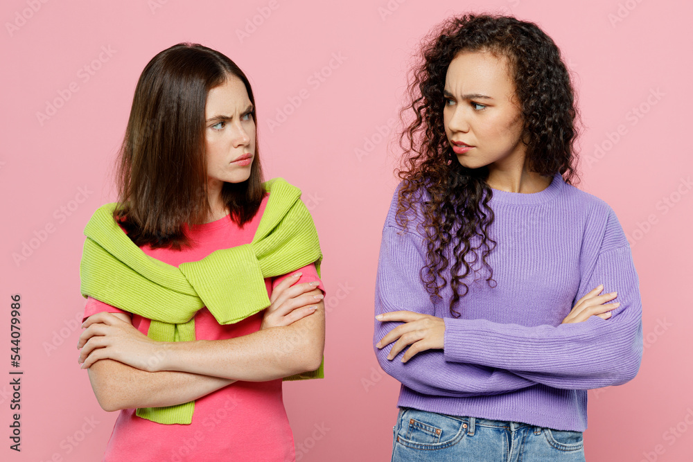 Young two friends sad offended pouted women 20s wearing green purple shirts look to each other hold hands crossed folded together isolated on pastel plain light pink color background studio portrait. Young two friends sad offended pouted women 20s wearing green purple shirts look to each other hold hands crossed folded together isolated on pastel plain light pink color background studio portrait.