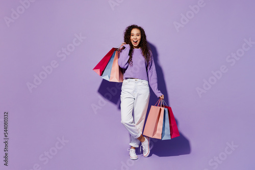 Full body happy surprised young woman 20s wears pullover hold in hand paper package bags after shopping look camera isolated on plain pastel light purple background. Black Friday sale buy day concept.