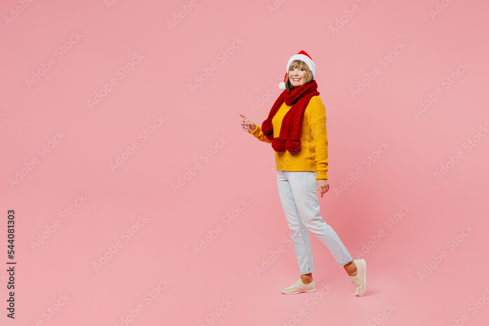 Full body side view fun elderly woman 50s years old wear yellow knitted sweater red scarf Santa hat posing point finger aside isolated on plain pink background. Happy New Year Christmas 2023 concept. Full body side view fun elderly woman 50s years old wear yellow knitted sweater red scarf Santa hat posing point finger aside isolated on plain pink background. Happy New Year Christmas 2023 concept.