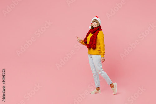Full body side view fun elderly woman 50s years old wear yellow knitted sweater red scarf Santa hat posing point finger aside isolated on plain pink background. Happy New Year Christmas 2023 concept.