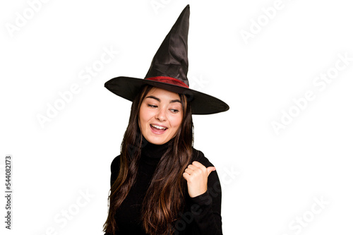 Young caucasian woman dressed as a witch isolated on green chroma background points with thumb finger away, laughing and carefree.