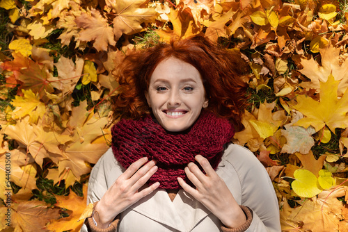 Smiling caucasian millennial woman with red hair in raincoat with scarf lies on ground with yellow leaves