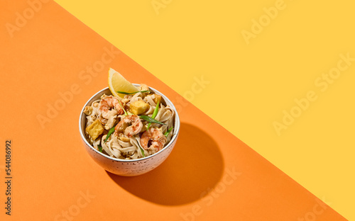 Asian street food - pad thai noodles with shrimp on coloured background. Pad thai udon with prawns and lemon in bowl on orange and yellow backdrop. Trendy concept udon noodles with seafood. photo