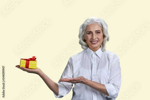 Beautiful smiling mature woman pointing at gift box on her palm. Pretty senior woman showing gift box and looking at camera isolated on yellow background.