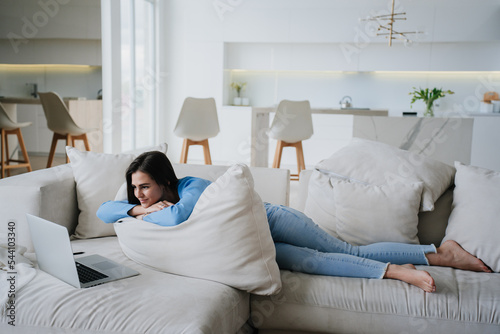 Young hispanic woman in blouse, jeans laying on sofa in front of laptop, leaning on a large pillow, watching  movie. Girl makes video call, education and remote work concept. Relax, leisure.