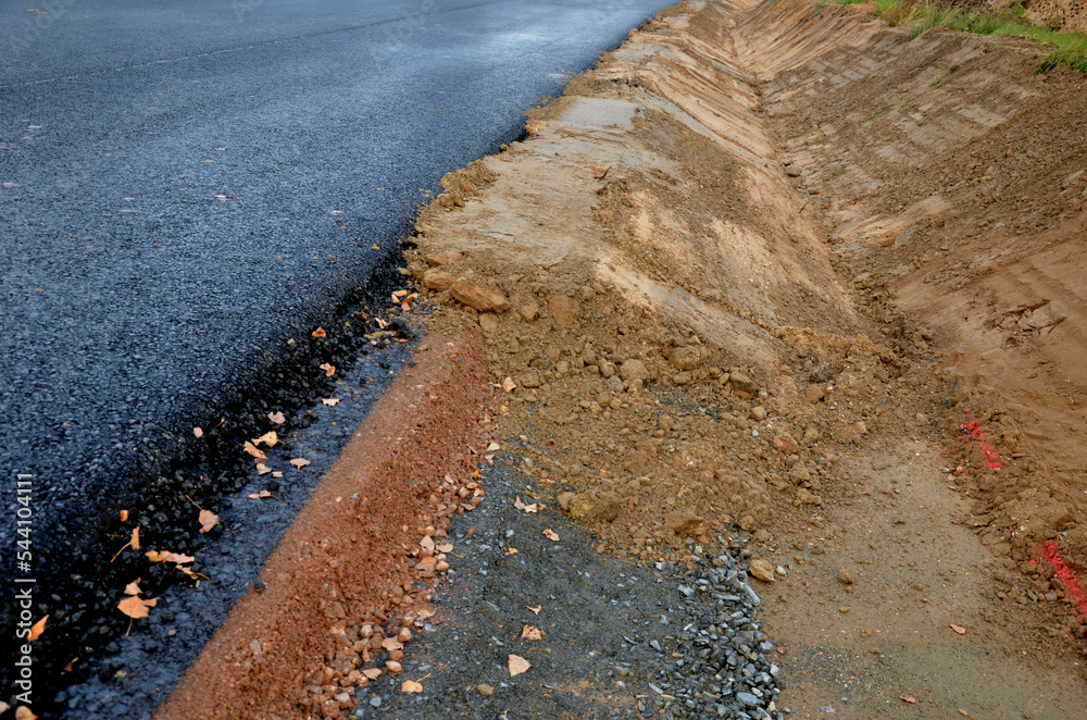 A newly built road, on the layer of which you can see successive layers of screed and asphalt coatings and penetration spraying. the edge is gradually shaped by earth at ditch, uncomplete, fall down 