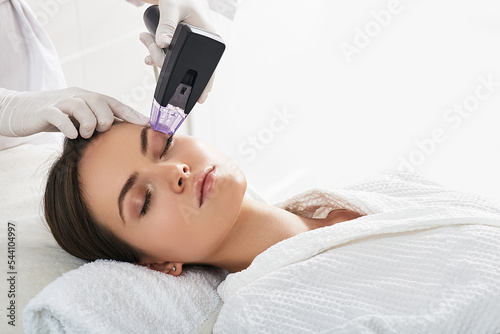 Brown haired woman receiving radiofrequency lifting procedure around eyes for her face skin rejuvenation at aesthetic cosmetology center