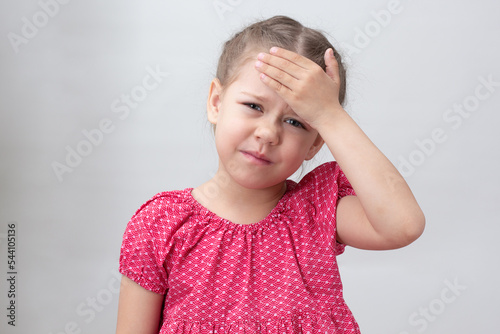 Child with headache holding hand on forehead on white background caucasian little girl 5-6 years in red looking at camera