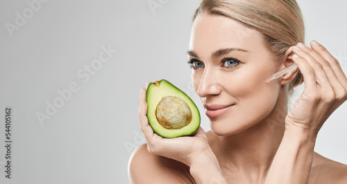 Caucasian woman applying avocado oil on her face with pipette dropper for nourishing and moisturizing skin and holds half of avocado, high quality. Facial skin care with avocado oil photo