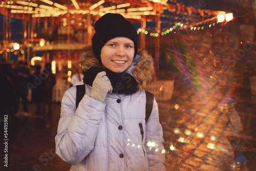 Outdoors on a festive square against the backdrop of a festival entertainment carousel  a happy teenager girl smiles broadly looking at the camera  space for text.