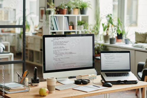 Workplace of modern IT support engineer with computer monitor and laptop with coded data on screens in openspace office