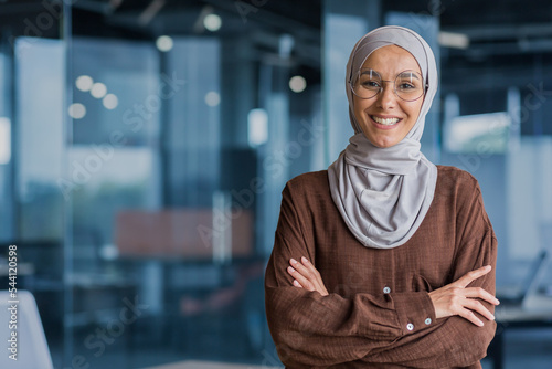 Bild auf Leinwand Portrait of successful and happy businesswoman in hijab, office worker smiling and looking at camera with crossed arms, working inside modern office