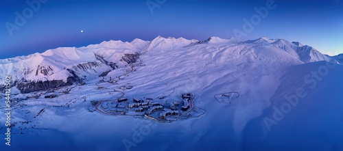 Wide aerial panorama of snowy mountain ridge on winter sunrise. Stunning mountains range covered with snow powder on ski resort at sunset. Moon above caucasus peaks skyline in the dusk at night.
