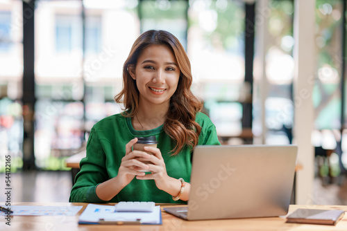 Beautiful Asian businesswoman sitting on her laptop happily working on her laptop and drinking coffee with a bright smile in the office and looking at the camera.