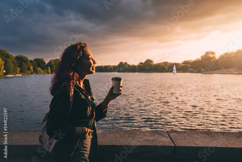 Photographie A student girl with a backpack drinks fragrant coffee near the embankment of Lak