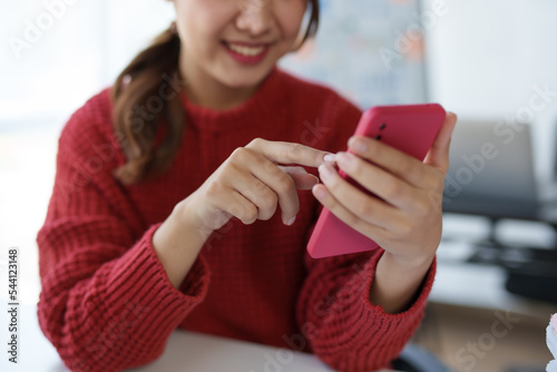 Smiling and charming Asian woman sit at her desk with a smartphone playing social media applications and transacting at her desk. photo