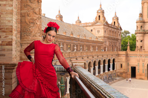Beautiful teenage woman dancing flamenco on the balcony of a square in Seville. She wears a red dress with ruffles with a lot of art. Flamenco cultural heritage of humanity.