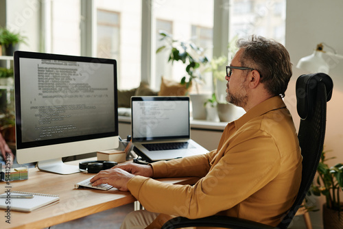 Serious mature software developer looking at code on computer screen while decoding data and carrying out working tasks in office