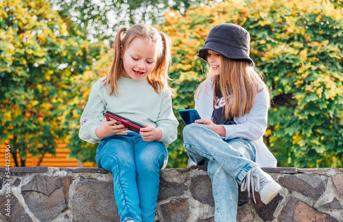 Two smiling little sister girls kids sitting and browsing their smartphone devices in the autumnal park. Careless young childhood time and a modern technology concept image.