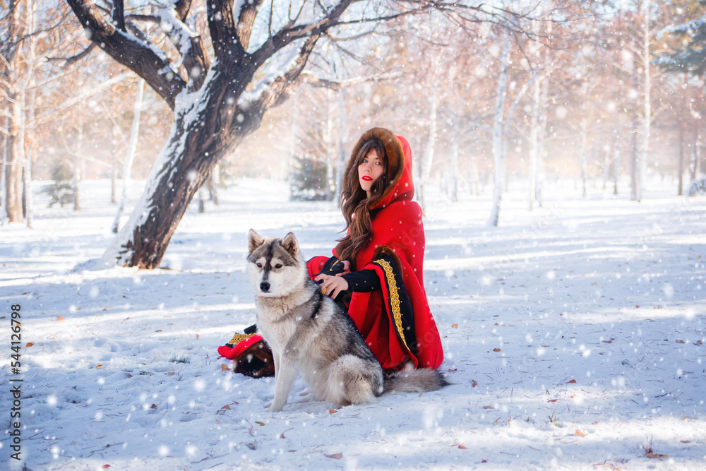 A young woman in a red hooded raincoat with a gray husky dog on the background of a winter forest and snowfall. A young woman in a red hooded raincoat with a gray husky dog on the background of a winter forest and snowfall.