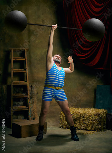 Vintage portrait of retro circus strongman wearing blue striped sports swimsuit training with barbell over dark circus backstage background. Art, fashion, style photo