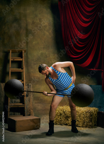 Vintage portrait of retro circus strongman wearing blue striped sports swimsuit training with barbell over dark circus backstage background. Art, fashion, style photo