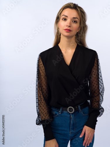 A beautiful, young, slender girl with long blonde hair on a white background in blue jeans and a stylish black blouse with transparent sleeves. Close-up
