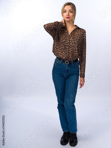 A beautiful, young, slender girl with long blonde hair on a white background in blue jeans and a stylish blouse with leopard spots. Full height