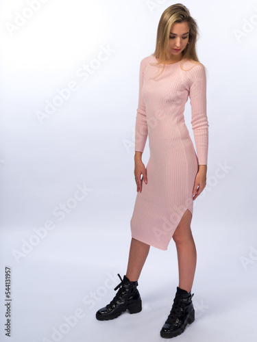 A beautiful, young, slender girl with long blonde hair on a white background. In a long pink knitted tight dress