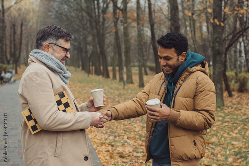 Friends, a senior and a young man walking and talking and drinking coffee together in the autumn park.. © dsheremeta
