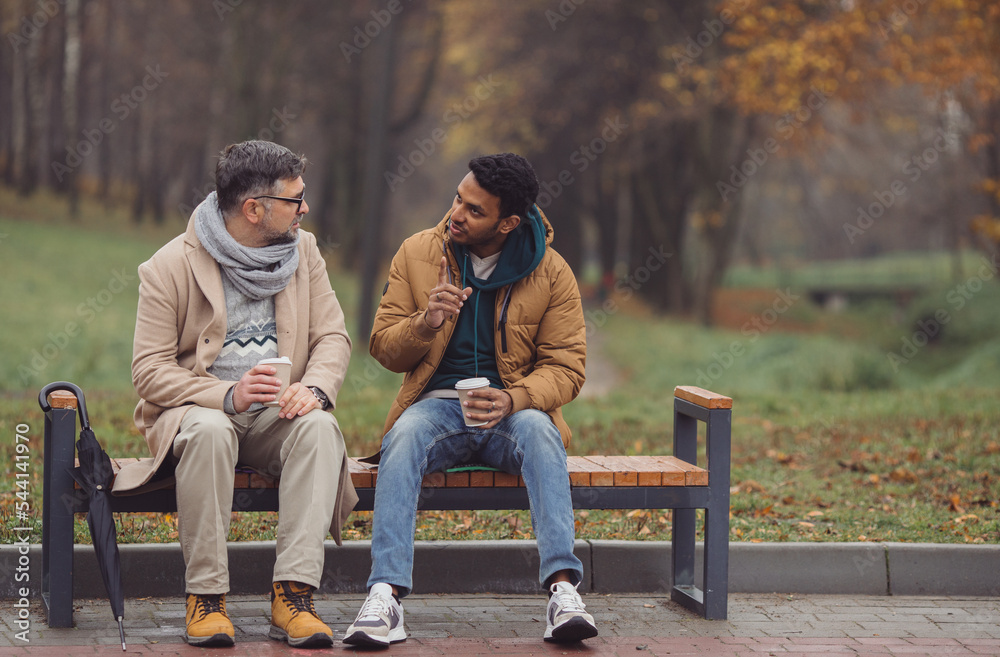 Friends, a senior and a young man sit in the park on a bench and talk in the autumn park. Friends, a senior and a young man sit in the park on a bench and talk in the autumn park.