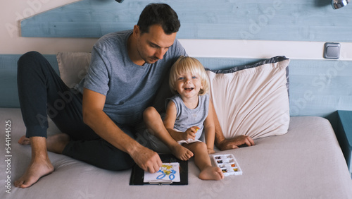 A small caucasian boy with his father on the bed draw with colored pencils.