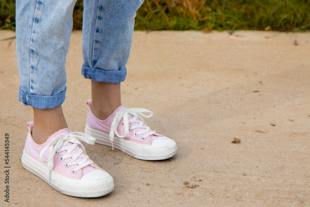 Naklejka premium A view of the legs of a teenage girl in pink sneakers and blue jeans against a background of yellow dry grass. The concept of clothing for modern teenagers, even in the cool season