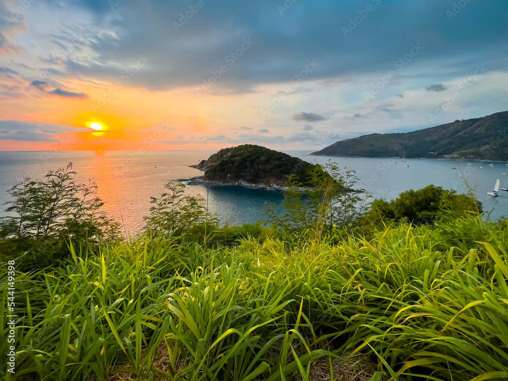Summer island sunset background. Beautiful tropical landscape aerial view. Colorful sky with orange sun over green grass meadow. Turquoise sea, yachts, ocean bay. Windmill Viewpoint, Phuket, Thailand