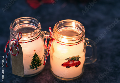 Lit Christmas candles of white paraffin in a glass, decorated with a car and a Christmas tree on a gray background.