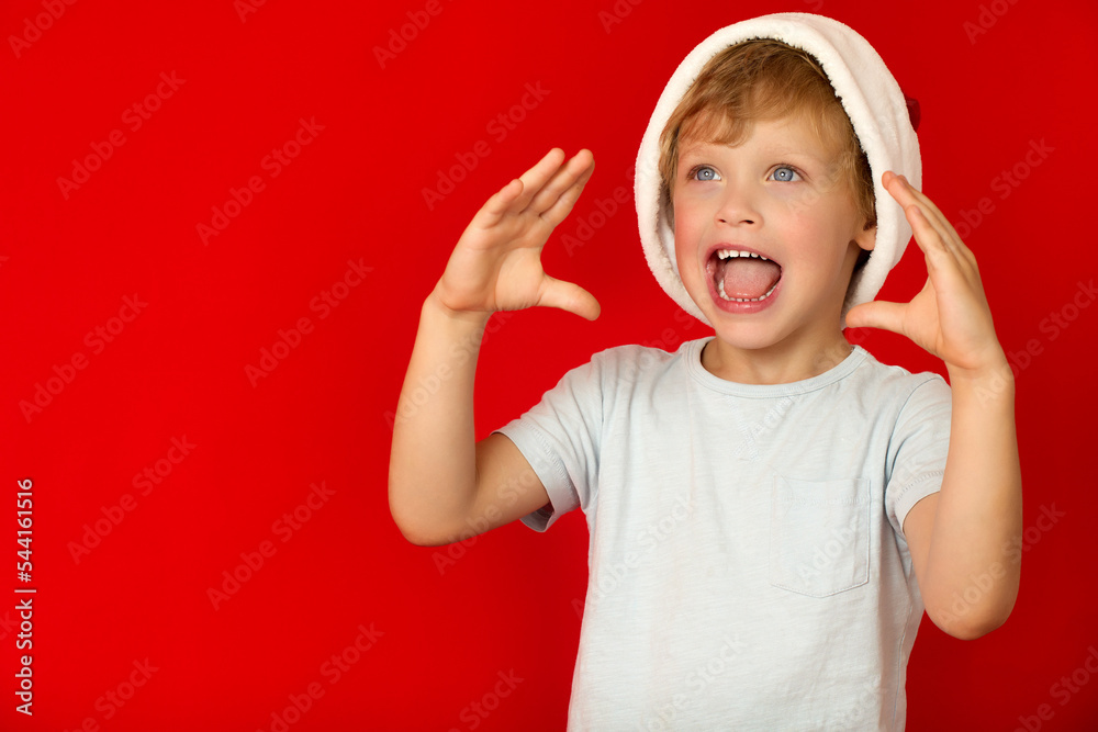 Screaming little boy with his mouth wide open on red background. Cute boy in red santa cap holds his hand near his open mouth, screams looking up Screaming little boy with his mouth wide open on red background. Cute boy in red santa cap holds his hand near his open mouth, screams looking up