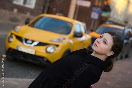 beautiful girl in the city. person in the street. a cute teenager in a black coat against a background of a blurred yellow car.