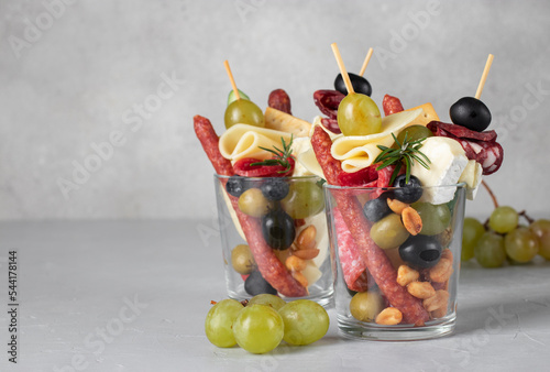 Jarcuterie in two glass includes various types of sausages, pickles, cheese, nuts, vegetables and fruits. Modern version of festive snack on light gray background, Copy space photo