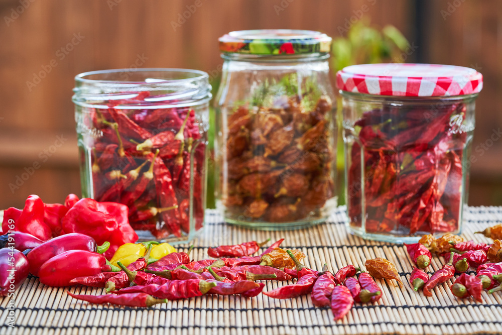 Preserving jars full of sun dried sweet and spicy red and yellow chilli peppers from organic agriculture and other fresh harvested riped peppers around them on the asian style bamboo mat outside. Preserving jars full of sun dried sweet and spicy red and yellow chilli peppers from organic agriculture and other fresh harvested riped peppers around them on the asian style bamboo mat outside.