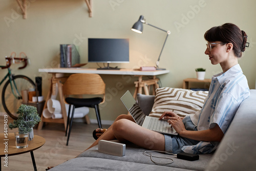Side view portrait of smiling young woman using laptop while relaxing on sofa in cozy home interior, copy space
