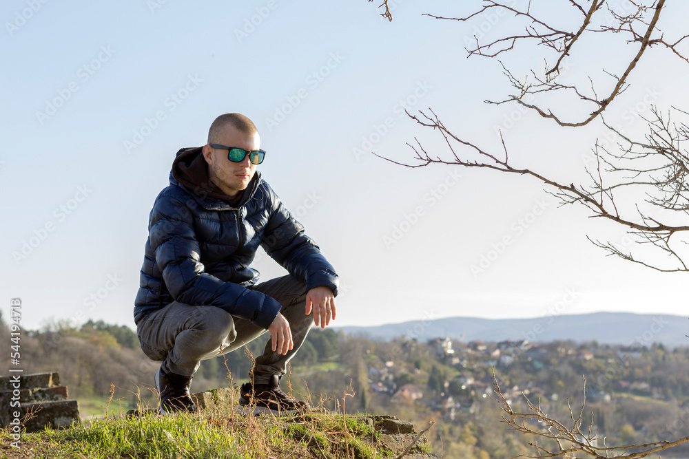 Stock photo of a young guy posing squatting in front of a city view on a winter sunny day.