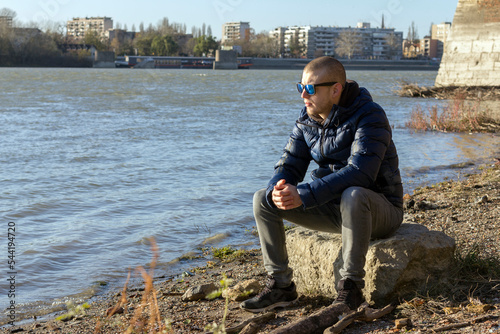Stock picture of a middle-aged man sitting on a coast by the river and looking at it thoughtfully, during a cold, sunny autumn day.