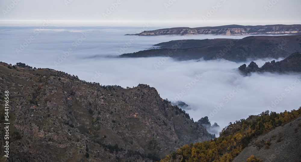 Rocky vertical cliffs of mountains protruding from above above the fog and clouds on an autumn day in the mountains of the Caucasus, hills with scree and yellowed vegetation Rocky vertical cliffs of mountains protruding from above above the fog and clouds on an autumn day in the mountains of the Caucasus, hills with scree and yellowed vegetation