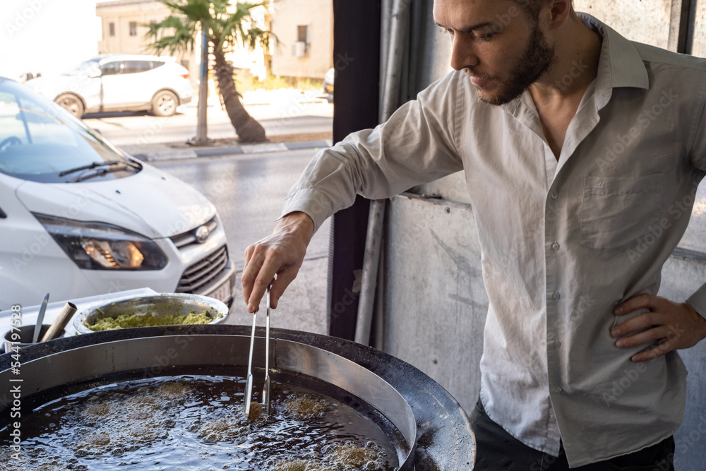 Fototapeta premium Young White Man in Grey Shirt Moves a Clamp and Fries Falafel Balls in a Giant Pan Filled with Boiling Oil in Middle Eastern Restaurant