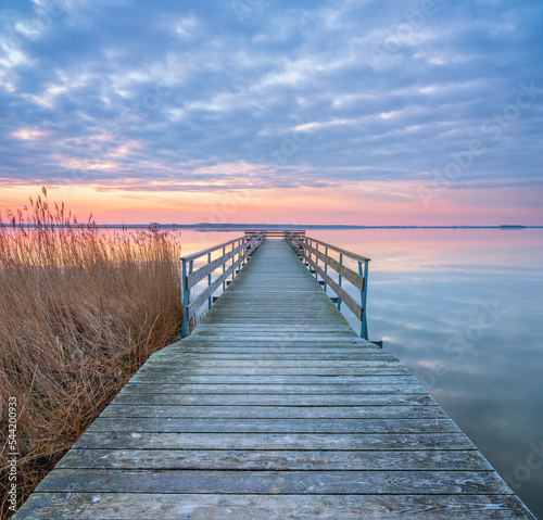 Calm Lake at Sunrise with Long Wooden Pier and Reeds under cloudy sky at sunrise © AVTG