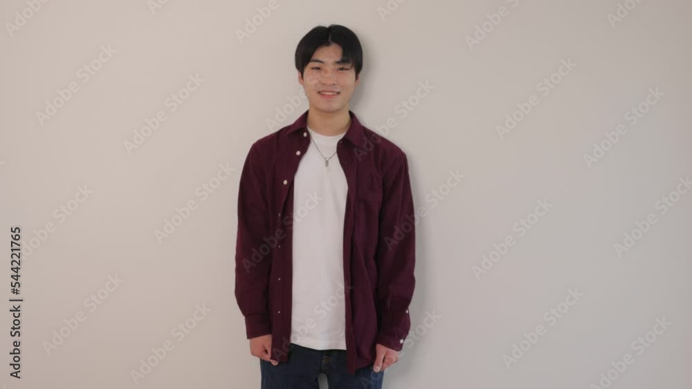 Asian boy standing still against the white wall and smiles. Wide shot.
