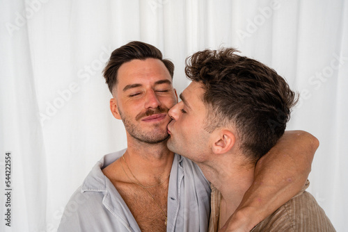 Gay couple kiss on chin with white curtain background