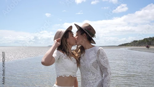 country lesbian brides laughing and kissing on beach while holding hats on wedding day