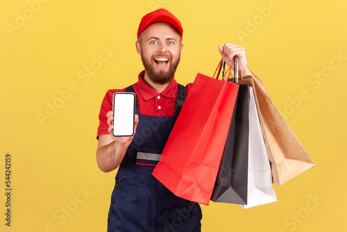 Portrait of happy smiling courier man holding cell phone and shopping bags, advertising online store on mobile device, good delivery application. Indoor studio shot isolated on yellow background.